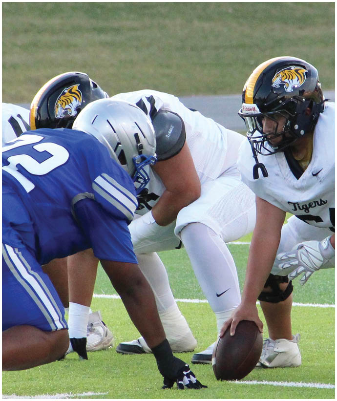 Two football players in white uniforms with tiger logos on their helmets are lined up against a player in a blue uniform and silver helmet on a grassy field. Description generated by AI