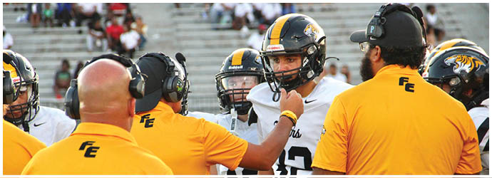 People wearing yellow shirts with headsets are interacting with two people in white football uniforms on a sports field. Description generated by AI