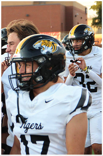 Two people wearing black helmets with a tiger logo and white sports jerseys are preparing for a football game outdoors near a brick building. Description generated by AI
