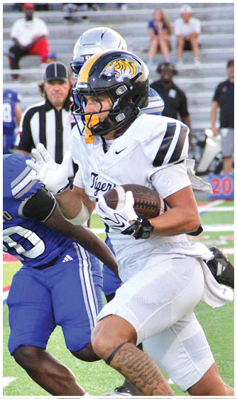A person in a white football uniform with a tiger logo on the helmet is running with the ball while being pursued by a person in a blue uniform on a football field. Description generated by AI