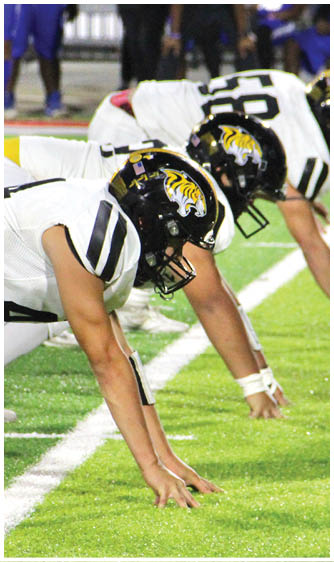 A person wearing a black football helmet with a yellow tiger logo and a white jersey with black stripes is leaning forward on a football field. Description generated by AI