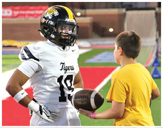 A person in a football uniform and helmet stands on a field facing another person in a yellow shirt holding a football. Description generated by AI