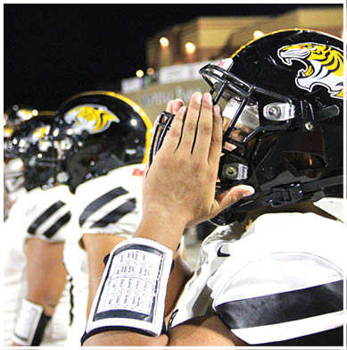 A person wearing a football helmet and white uniform is holding their hands together near their face, with another person in a similar uniform and helmet visible in the background. Description generated by AI