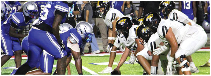 Two opposing American football teams in blue and white uniforms are lined up facing each other on the field with spectators in the background. Description generated by AI