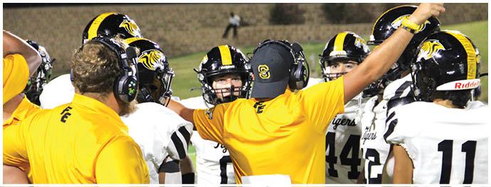People wearing football helmets and uniforms are gathered on a field with two adults in yellow shirts appearing to coach or instruct them. Description generated by AI