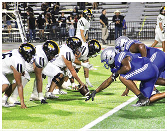 Two opposing American football teams in white and blue uniforms are lined up facing each other on a green field, ready for the snap. Description generated by AI