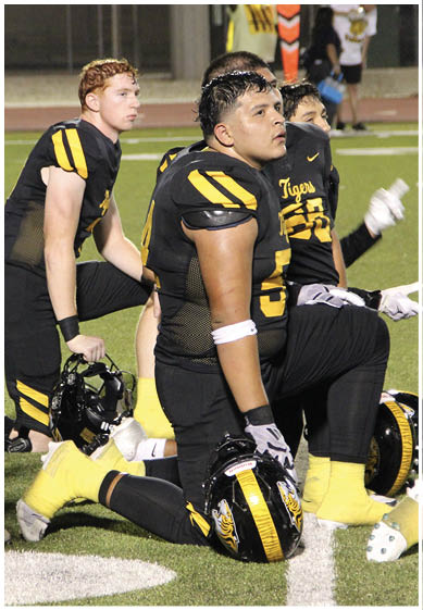 A group of people wearing black and yellow football uniforms with the word Tigers are sitting on the field, appearing focused and attentive. Description generated by AI
