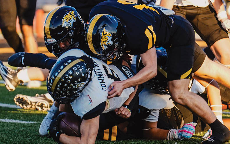 Several people wearing football helmets and uniforms are engaged in a tackle on a grassy field during a game. Description generated by AI