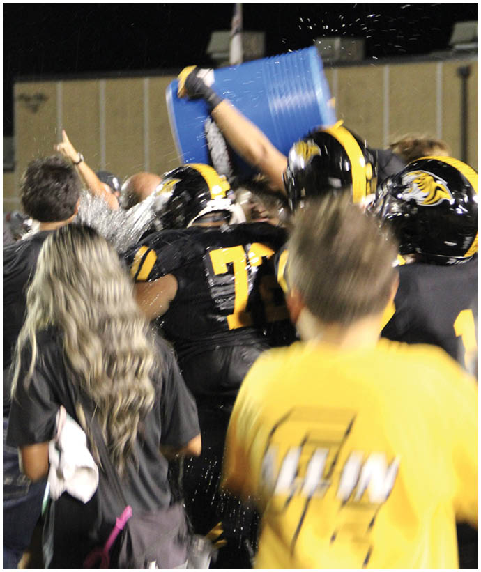 People in football uniforms celebrate as one person pours water from a large blue container over the group. Description generated by AI
