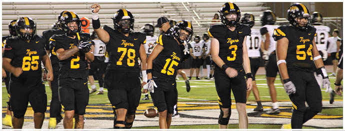Six people in black and yellow football uniforms walk together on a field with a crowd and other players in the background. Description generated by AI