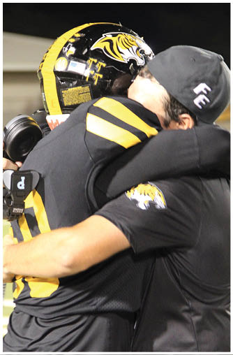 Two people in black and yellow sports uniforms embrace on a field, with one wearing a helmet featuring a tiger logo. Description generated by AI