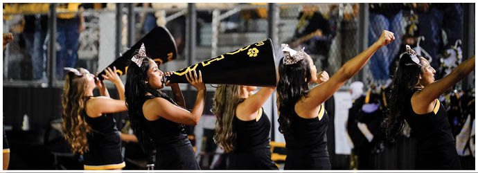 A group of people dressed in black cheerleading uniforms with bows in their hair are performing with one person using a megaphone at an outdoor event. Description generated by AI