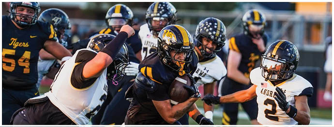 A group of people in football uniforms are actively playing on a field with one person holding the ball while others attempt to block or tackle. Description generated by AI