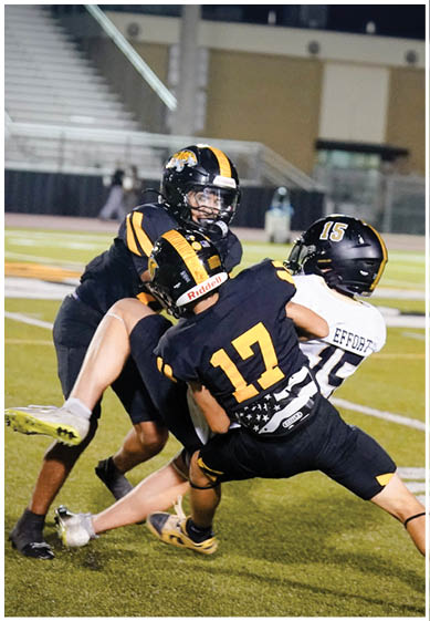Three football players in black and white uniforms are engaged in a tackle on a field with empty bleachers in the background. Description generated by AI