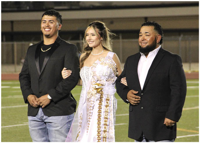 Three people stand arm in arm on a sports field at night, with the person in the center wearing a white dress and a large decorative sash while the others wear dark jackets and jeans. Description generated by AI