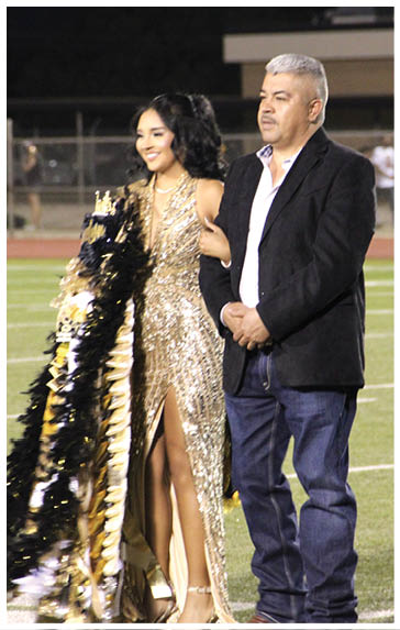 A person in a sparkling gold dress with a high slit stands arm-in-arm with another person wearing a black blazer and jeans on a sports field at night. Description generated by AI