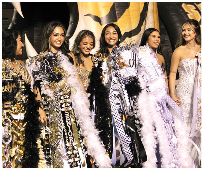 A group of people dressed in formal attire with decorated sashes and boas are standing closely together and smiling at an event. Description generated by AI