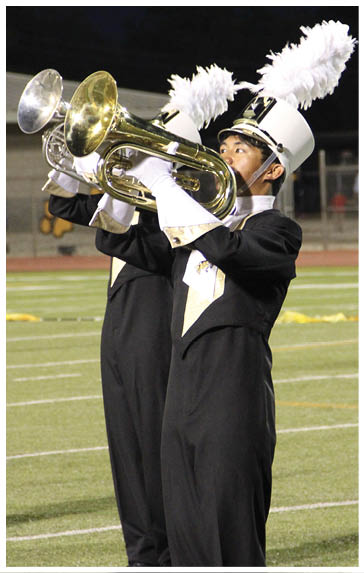 People in marching band uniforms with white plumed hats are playing brass instruments on a sports field at night. Description generated by AI