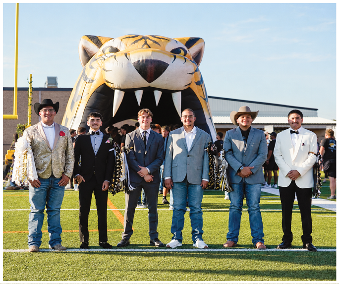 Six adults dressed in formal and semi-formal attire stand on a sports field in front of a large inflatable tiger head with its mouth open. Description generated by AI