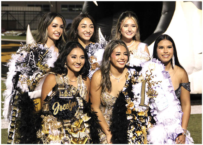 A group of six smiling people dressed in decorated formal attire with sashes and accessories pose together outdoors at night. Description generated by AI
