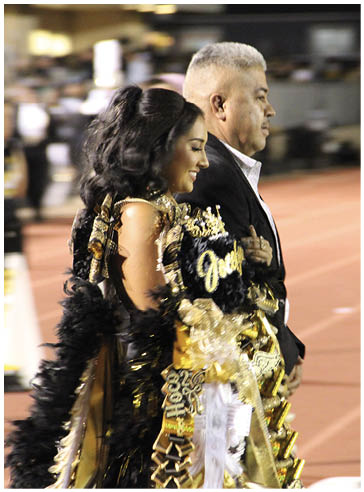 An adult with styled long hair wearing a decorated sash and formal attire is walking arm-in-arm with a senior in a suit on a track field. Description generated by AI