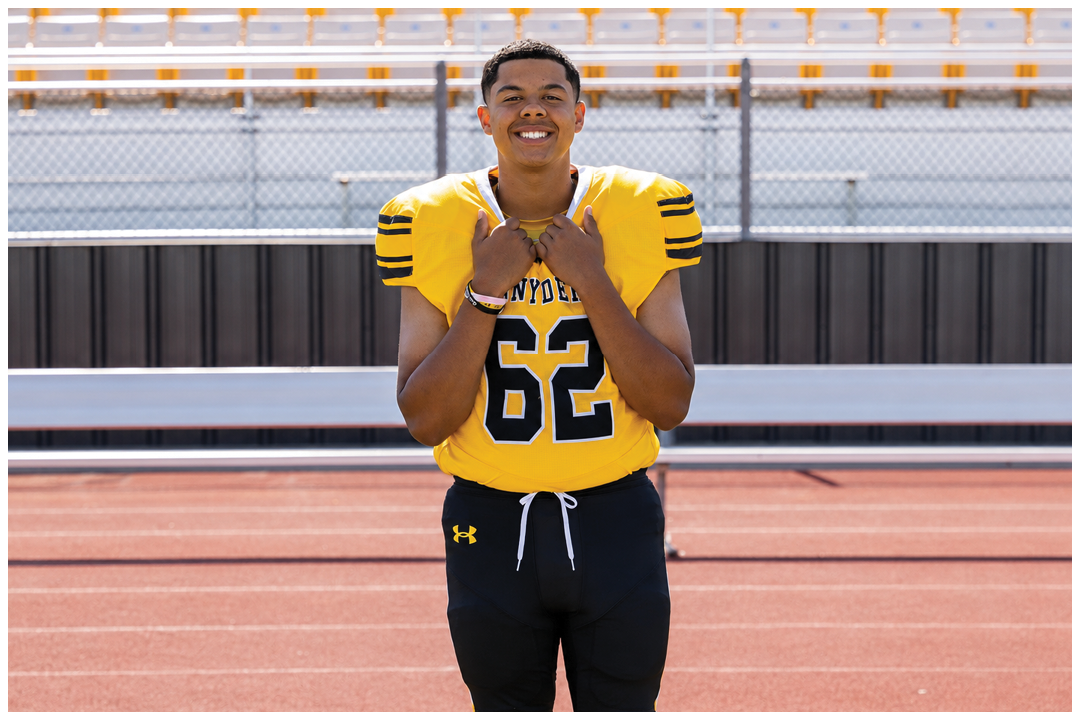 A person wearing a yellow football jersey with the number 62 and black shorts is standing on a sports field with a running track and bleachers in the background. Description generated by AI