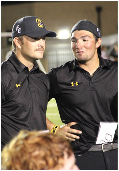 Two adults wearing black Under Armour shirts and caps are standing close together on a sports field, with one person’s arm around the other. Description generated by AI