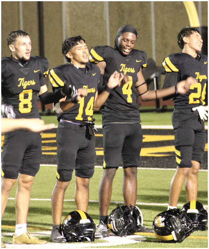 Four people wearing black and yellow football uniforms with the word Tigers stand on a field holding hands and appear to be celebrating or cheering. Description generated by AI