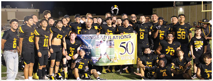A group of people wearing black and yellow sports uniforms are gathered on a football field at night holding a banner celebrating a coach's 50 wins. Description generated by AI