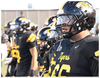People wearing black and yellow football helmets and uniforms are lined up, with one person in the foreground looking focused. Description generated by AI