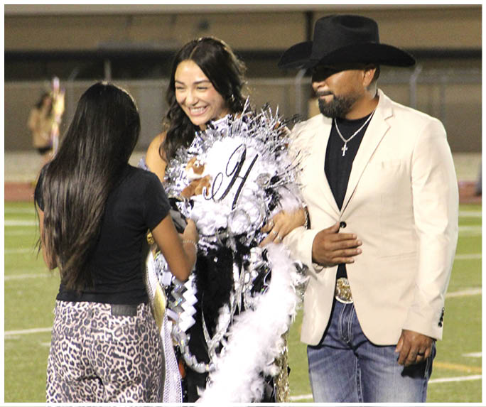 Three people are interacting happily, with one person wearing a decorative sash and another wearing a light-colored jacket and black hat. Description generated by AI