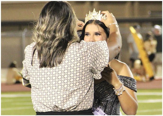 A person with long hair is placing a crown on another person wearing a sparkly off-shoulder dress at an outdoor event. Description generated by AI