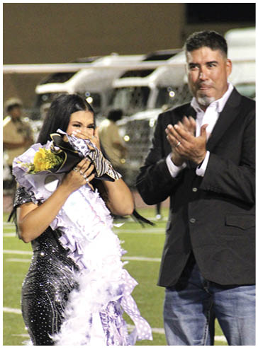 A person wearing a sparkly dress holds flowers and a large decorative sash while another person in a blazer and jeans claps beside them on a sports field. Description generated by AI