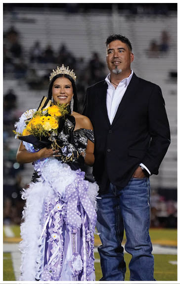 A person wearing a crown and a decorated sash holds a bouquet of yellow flowers while standing next to another person dressed in a blazer and jeans on a sports field with bleachers in the background. Description generated by AI