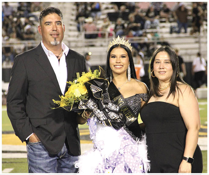 Three people stand together on a sports field with one person in the center wearing a crown, sash, and holding a bouquet of flowers, while the other two stand on either side dressed in formal attire. Description generated by AI