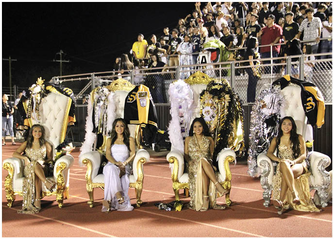 Four people dressed in elegant gowns are seated on ornate golden and white chairs on a track field with decorated jackets and a crowd in the background. Description generated by AI
