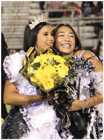 Two people smiling and hugging each other while holding a bouquet of yellow flowers and wearing formal attire with one person wearing a crown and sash. Description generated by AI