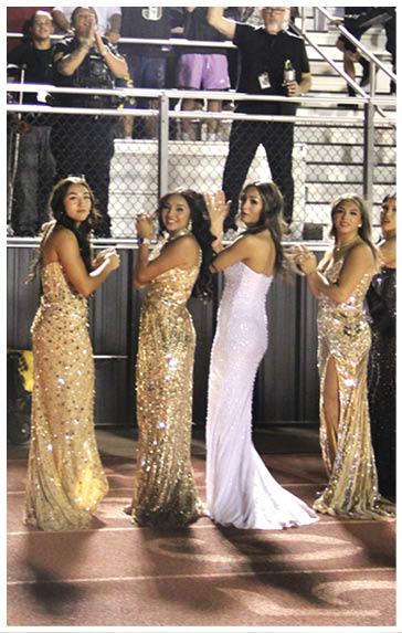 Four adults dressed in elegant, sparkling gowns stand in a row on a track field with a crowd of people cheering behind a fence. Description generated by AI