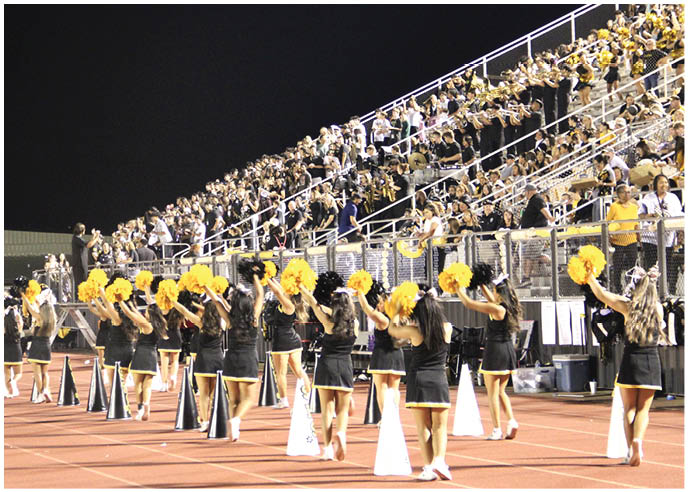 A group of cheerleaders in black outfits with yellow pom-poms perform on a track in front of a large crowd of people seated in bleachers at night. Description generated by AI