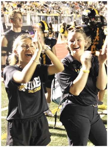Two adults wearing black sports-themed shirts are enthusiastically clapping and cheering at an outdoor event with a crowd and players in the background. Description generated by AI