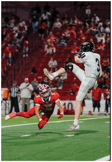 A person in a white football uniform is kicking the ball while another person in a red football uniform dives toward them on a football field with a crowd in the background. Description generated by AI