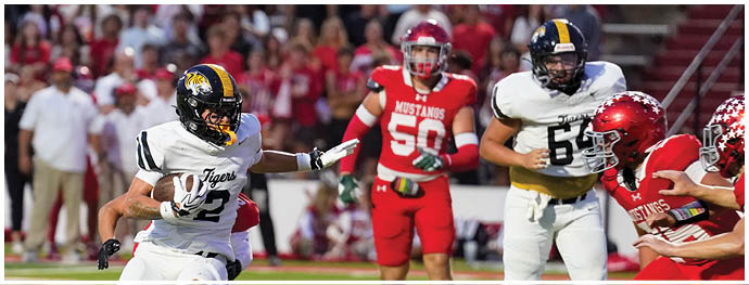 A person in a white football uniform with a ball is running on a field while several people in red uniforms attempt to block or tackle them. Description generated by AI