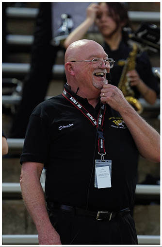 A senior person wearing glasses and a black polo shirt with a “Director" label is smiling and holding a microphone headset while standing outdoors near bleachers. Description generated by AI