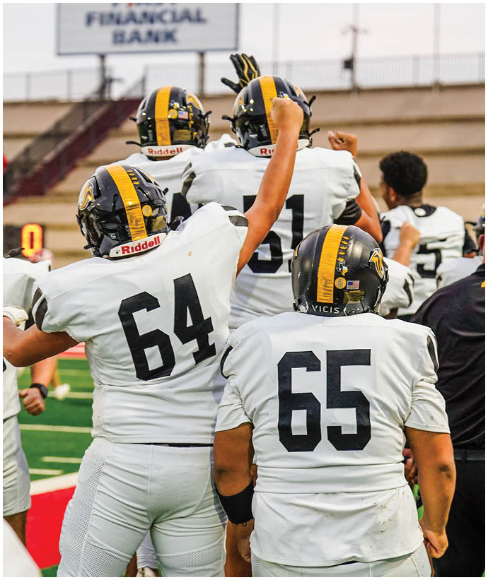 Four football players in white uniforms with black and yellow helmets are raising their fists on a field. Description generated by AI