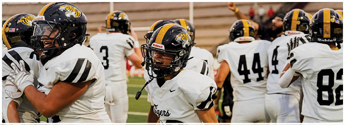 Several people wearing white football uniforms with black and yellow helmets are standing on a field, with one person in the center facing forward and others facing away. Description generated by AI