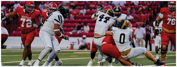 People in football uniforms are engaged in a tackle during a game with a crowd in the background. Description generated by AI