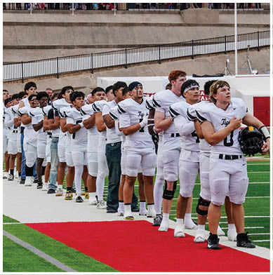 A line of people in white football uniforms stand on a field with their right hands over their hearts. Description generated by AI