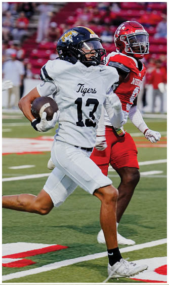 Two people in football uniforms are on a field with one wearing a white jersey labeled “Tigers" and the other in a red jersey, both appearing to be engaged in a game. Description generated by AI