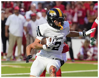 A person in a white football uniform with a tiger logo is running with the ball while players in red and white uniforms are chasing on a football field. Description generated by AI