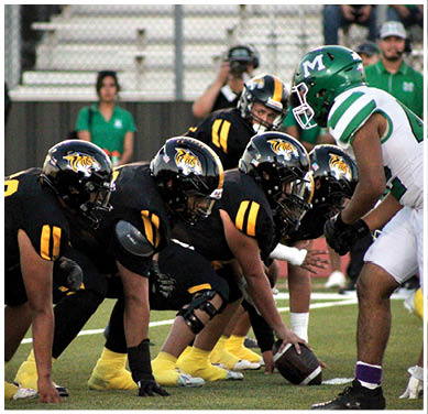 A group of football players in black and yellow uniforms are lined up on the field facing a player in a green and white uniform during a game. Description generated by AI